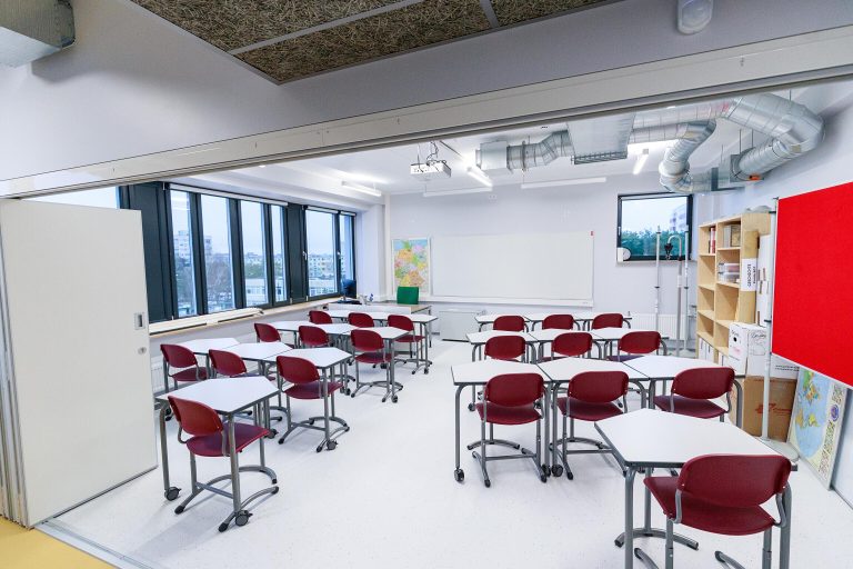 Empty classroom with red chairs and white desks arranged for students.