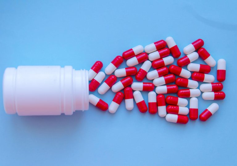 Open white medication bottle with red and white capsules scattered on a table.